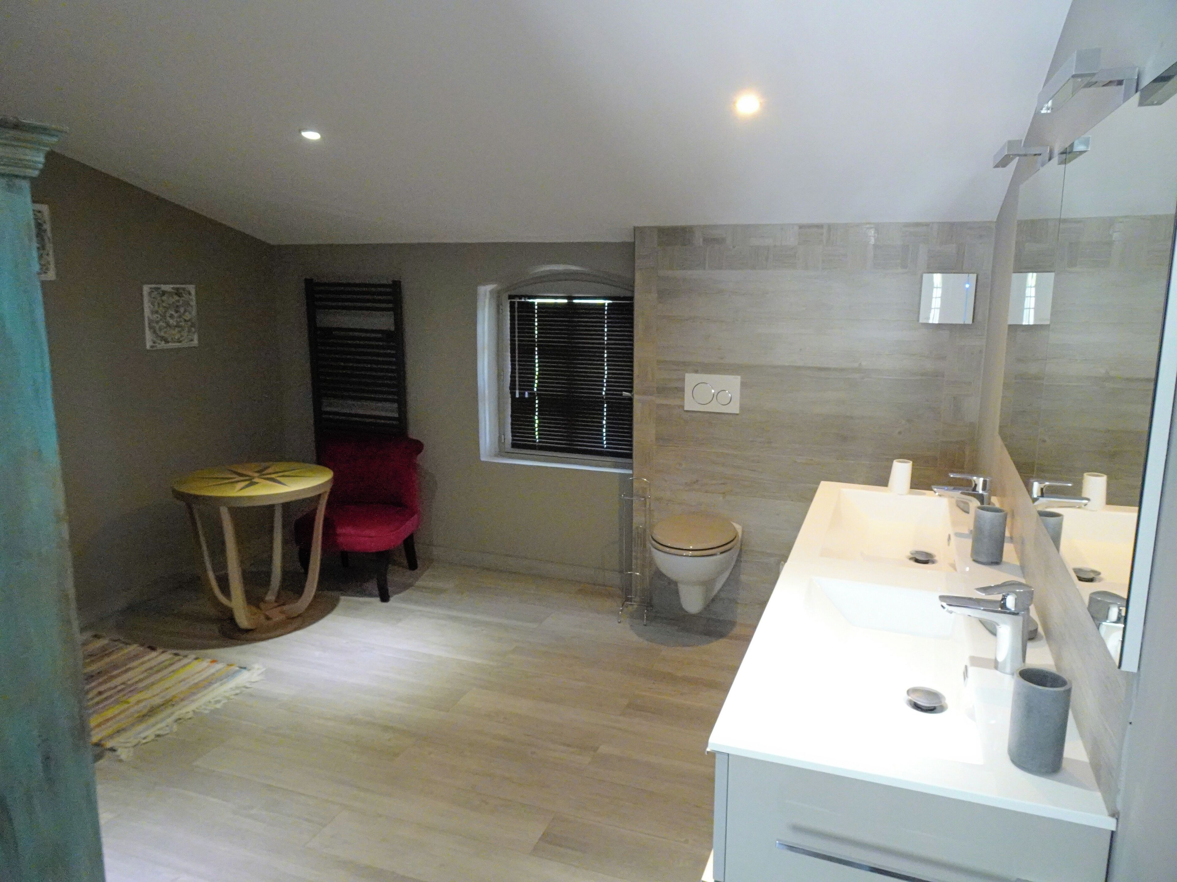 A modern bathroom with a large mirror, white sink, and a toilet. There is a small round table with a chair and a window with blinds.