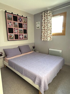 A bedroom with a bed, a nightstand, a wall-mounted quilt, and a window with curtains.
