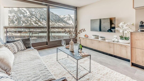 Ein modernes Wohnzimmer mit großem Fenster, einer Couch, einem Couchtisch und einer Küchenzeile mit Fernseher.