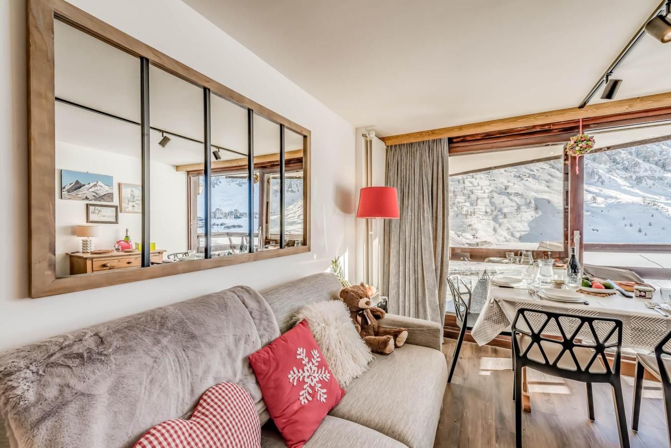 A cozy living room with a large mirror, a sofa with decorative pillows, a teddy bear, and a dining area with a table set for two.