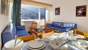 Ein gemütliches Zimmer mit einer Couch, einem Esstisch gedeckt mit Tellern und Besteck sowie Bergblick durch ein Fenster.