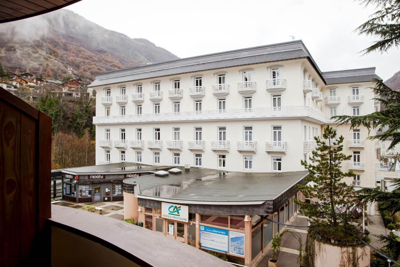 Un hotel de varios pisos con balcones, un espacio al aire libre cubierto y un paisaje montañoso al fondo.