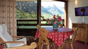 A room with a wooden table, chairs, a TV, and a large window with a mountain view.
