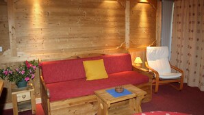A cozy room with a wooden wall, a red sofa with a yellow pillow, a wooden chair, and a small table with a lamp.