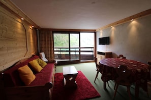 A room with a wooden wall, a sofa with cushions, a table with chairs, a television, and a large window with a view.