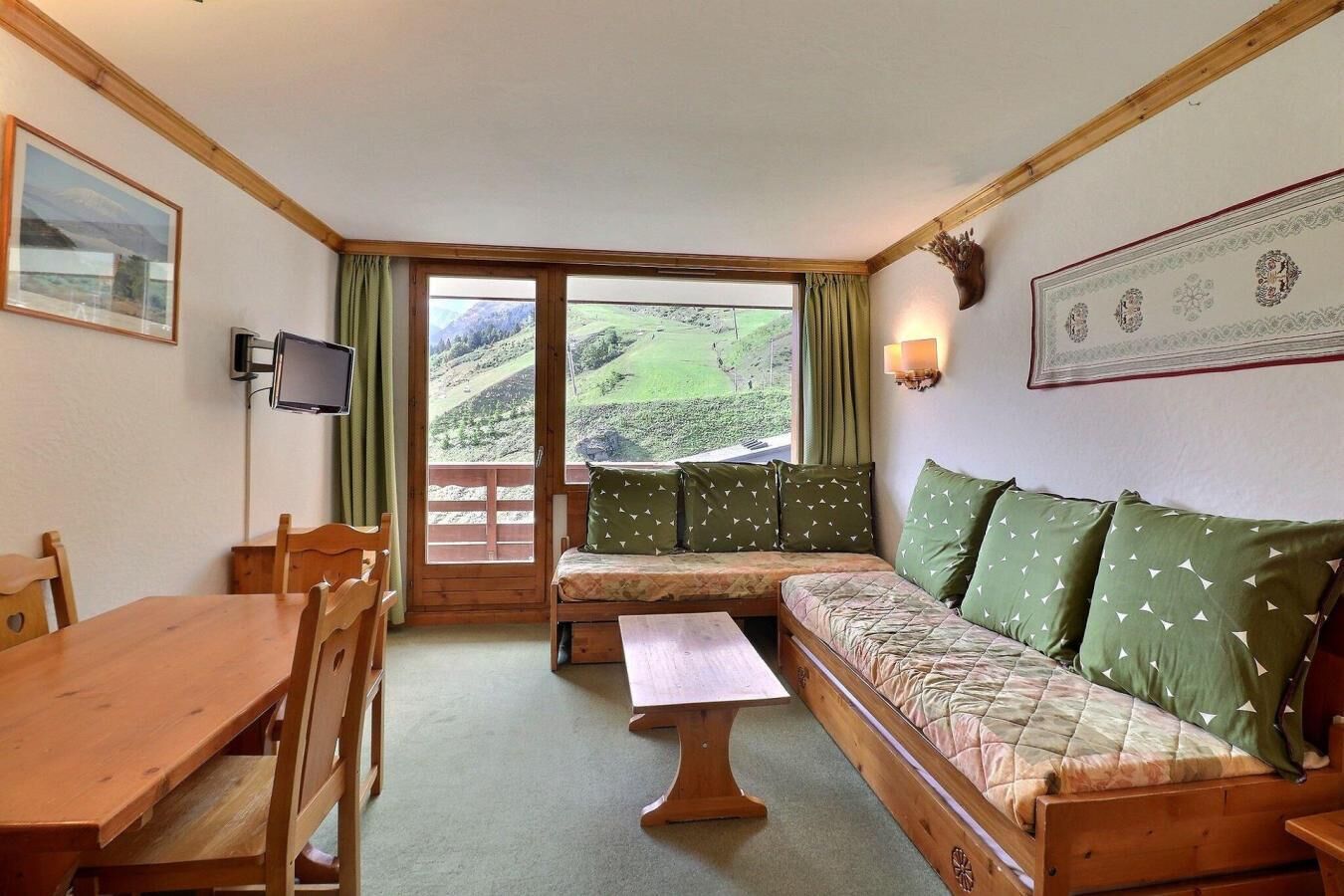 A cozy room with a wooden table, a sofa with patterned cushions, a television, and a large window offering a view of a green landscape.