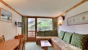A cozy room with a wooden table, a sofa with patterned cushions, a television, and a large window offering a view of a green landscape. - 10m from ski slopes, 6th floor, balcony, 40m², Méribel-Mottaret (Méribel - Les Allues)