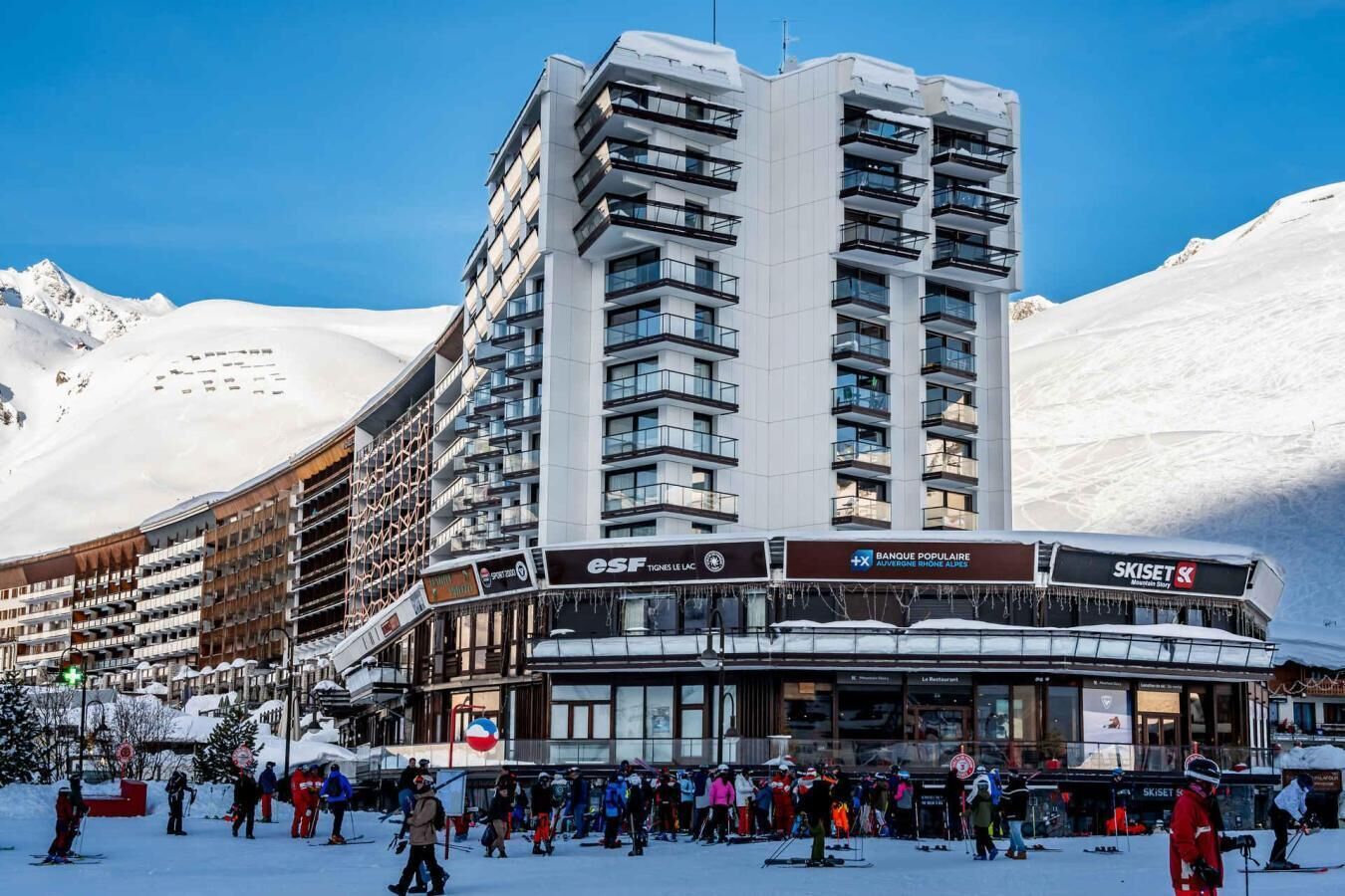 Un hotel moderno con diversi piani, negozi di articoli da sci e una stazione sciistica sullo sfondo.