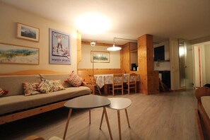 A cozy living room with a sofa, a dining table, and chairs. There are framed pictures on the wall and a lamp hanging above the table.