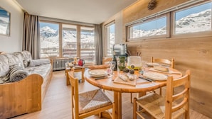 A cozy room with a wooden dining table, chairs, a sofa, and large windows offering a mountain view.