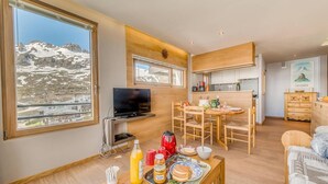 A modern living room with a large window, a flat-screen TV, wooden furniture, and a dining area with a table set for breakfast.