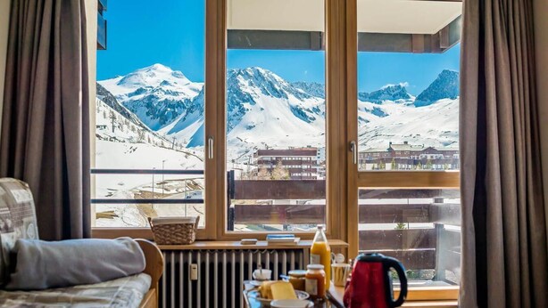 A cozy room with a view of snow-covered mountains, a wooden table with breakfast items, and a comfortable sofa.
