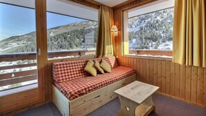 A cozy cabin room with a checkered sofa, a small wooden table, and large windows offering a view of snow-covered mountains. - 10m from ski slopes, 2nd floor, balcony, 33m², Méribel-Mottaret (Méribel - Les Allues)
