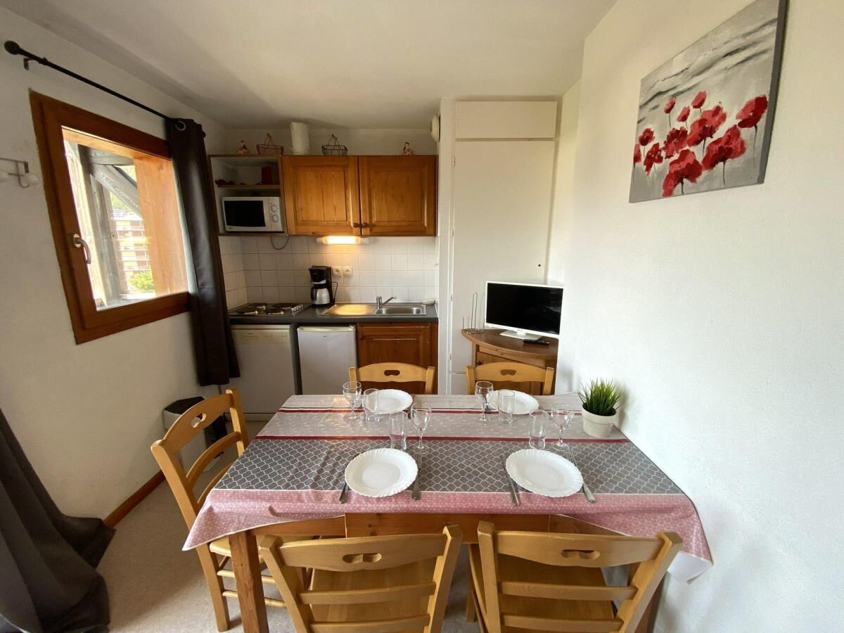 Un coin repas avec une table dressée pour quatre personnes, des chaises en bois, un téléviseur posé sur un meuble et une cuisine en arrière-plan.