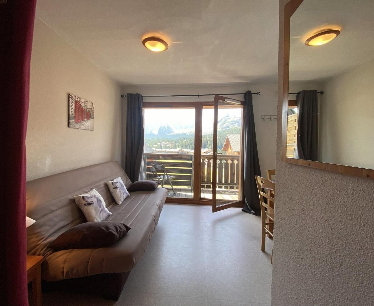 A room with a sofa, a chair, a balcony with a view, and a wooden floor.