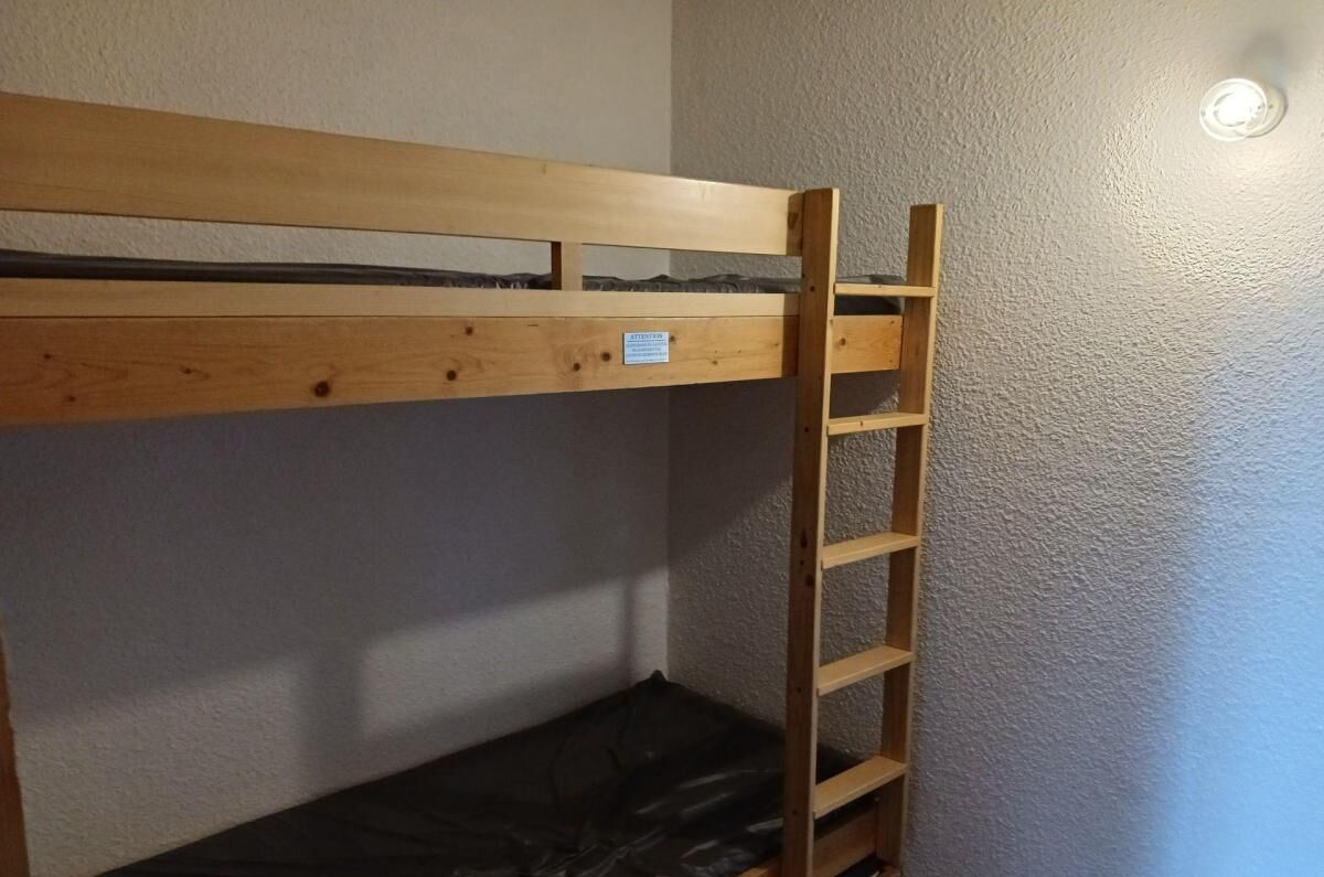 A bunk bed with a wooden ladder and a small shelf, against a textured wall with a light fixture.