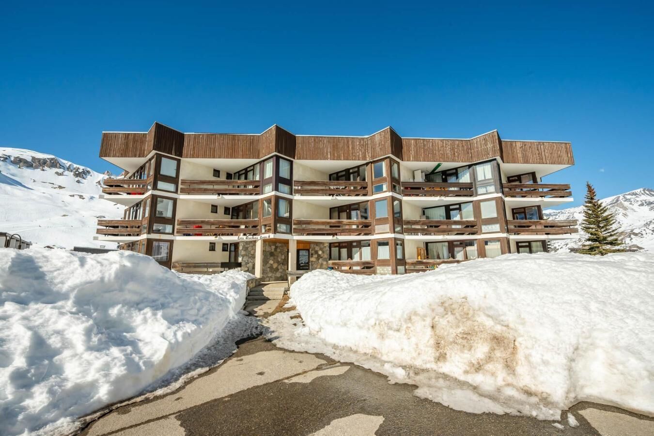 A multi-story hotel with balconies, a stone facade, and a snowy mountain backdrop.