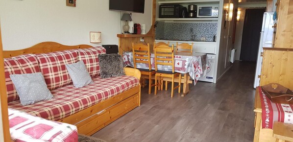A cozy living room with a plaid sofa, wooden chairs, a small table, and a kitchenette in the background.