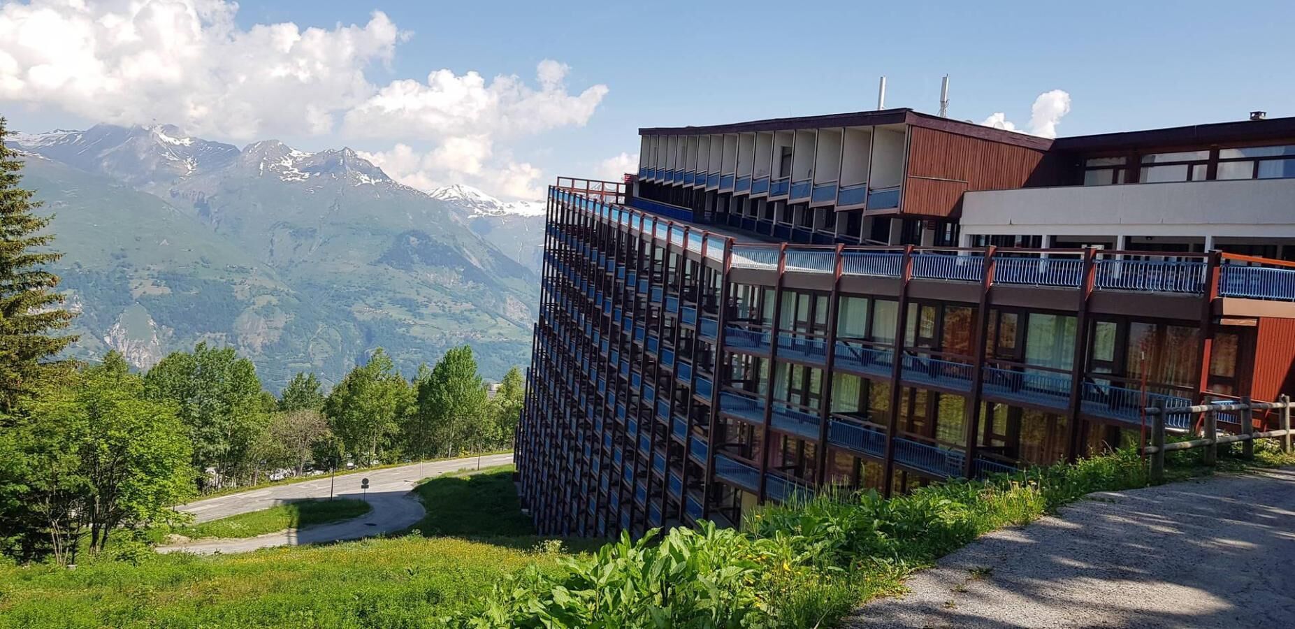 A modern building with multiple balconies, surrounded by lush greenery and mountains in the background.
