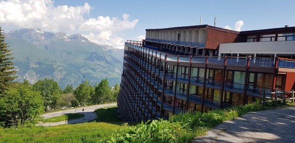 A modern building with multiple balconies, surrounded by lush greenery and mountains in the background.