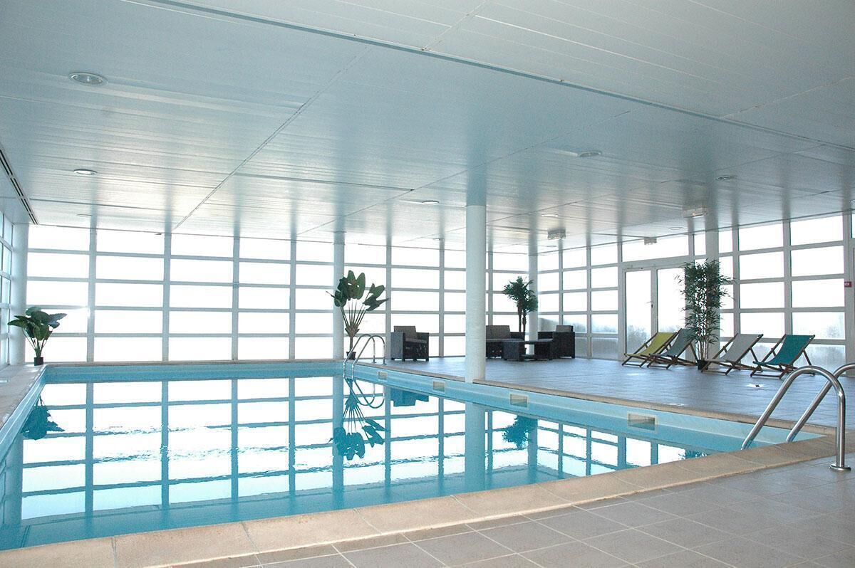 An indoor swimming pool with clear glass walls, a tiled floor, and several lounge chairs and potted plants around the pool area.
