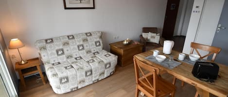 A cozy living room with a patterned sofa, a wooden dining table with chairs, a small side table, and a framed picture on the wall.