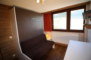 A compact room with a dark-colored sofa, a small table, a window with red curtains, and a TV mounted on the wall.