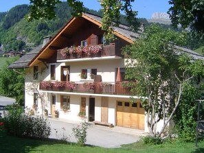 Eine zweistöckige Chalet mit Balkon, umgeben von viel Grün und einer Bergkulisse.