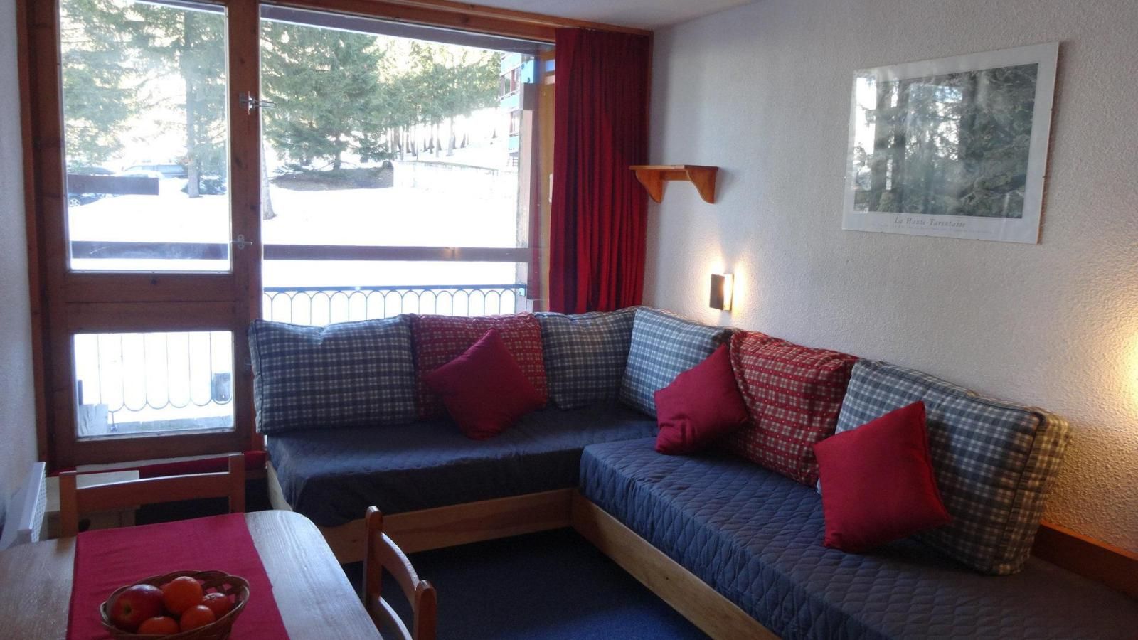 Ein gemütliches Zimmer mit einem blauen Sofa, roten Kissen, einem Holztisch mit roter Tischdecke und einem Fenster mit Blick auf Bäume und ein Gebäude.