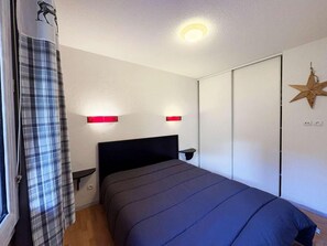 Une chambre à coucher avec un lit, une table de chevet, une lampe fixée au mur, une décoration en forme d’étoile et une fenêtre avec un rideau à carreaux.