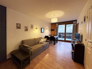 A cozy living room with a sofa, a small table, and a TV on a stand. There is a dining area with a table and chairs, and a balcony with a view.