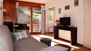 A cozy living room with a sofa, a television on a stand, and a view of a forested area through large windows. - Ski-in/ski-out & foot of the slopes, 2nd floor, view swimming pool, swimming pool, sauna, hammam (Les Orres)