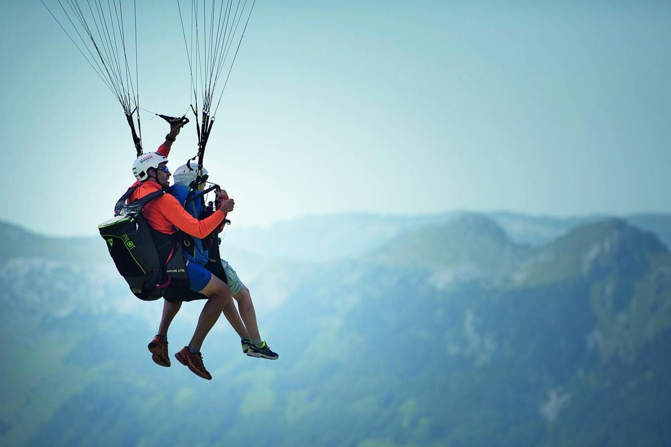 Two people paragliding with a mountainous landscape in the background.