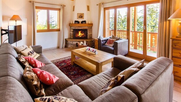 A cozy living room with a fireplace, a sofa set, and large windows offering a view of the outdoors.