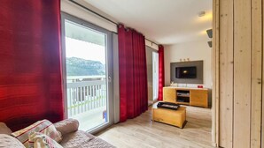 A living room with a sliding glass door, a flat-screen TV, wooden furniture, and a balcony with a view of a hilly landscape. - 100m from shops, 2nd floor, view mountain, tv, 30m², Flaine (Flaine)