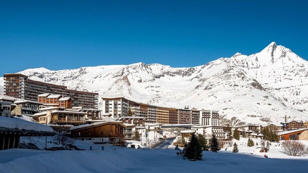 Ein verschneiter Ferienort mit mehreren Gebäuden, darunter ein großes Hotel mit Balkonen und einer erkennbaren Seilbahn.