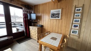 Ein gemütliches Zimmer mit einem Holztisch und Stühlen, einem Schminktisch, einem Fernseher und einer Wand mit gerahmten Bildern.
