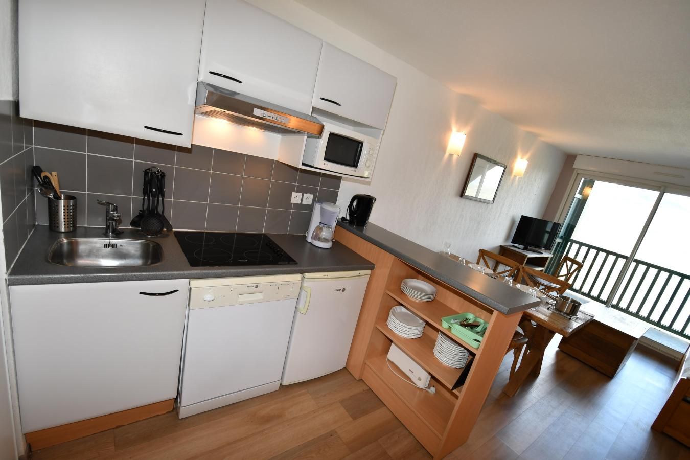 A modern kitchen with a built-in microwave, stove, and dishwasher. There is a wooden dining area with a table and chairs, and a balcony with a railing.