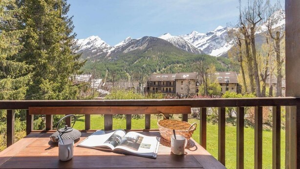 A wooden table with a book, a mug, and a basket on a balcony overlooking snow-capped mountains and a village. - 300m from resort center, 1st floor, balcony, tv, 33m², Serre Chevalier (Le Monêtier-les-Bains)