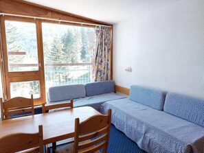 A room with a wooden table and chairs, a sofa with patterned cushions, and a large window with a view of trees and buildings.