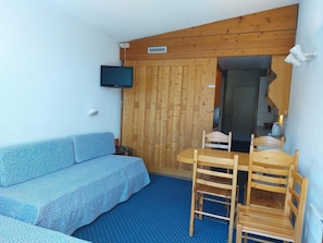 Um quarto pequeno com teto de madeira, um sofá azul, uma mesa de madeira com cadeiras e uma televisão fixada na parede.