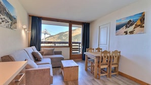 A cozy room with a sofa, a dining table with chairs, a small wooden cabinet, and a large window offering a mountain view. - 10m from ski slopes, 1st floor, balcony, 33m², Méribel-Mottaret (Méribel - Les Allues)
