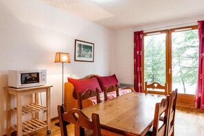 A cozy room with a wooden dining table, chairs, a sofa with red cushions, a standing lamp, a framed picture on the wall, and a window with curtains. - Swimming pool, terrace, parking, tv, ski locker, 30m², Super Besse (Super Besse)