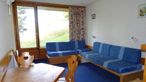 Una habitación con una mesa y sillas de madera, un sofá azul y una ventana con vistas a zonas verdes.