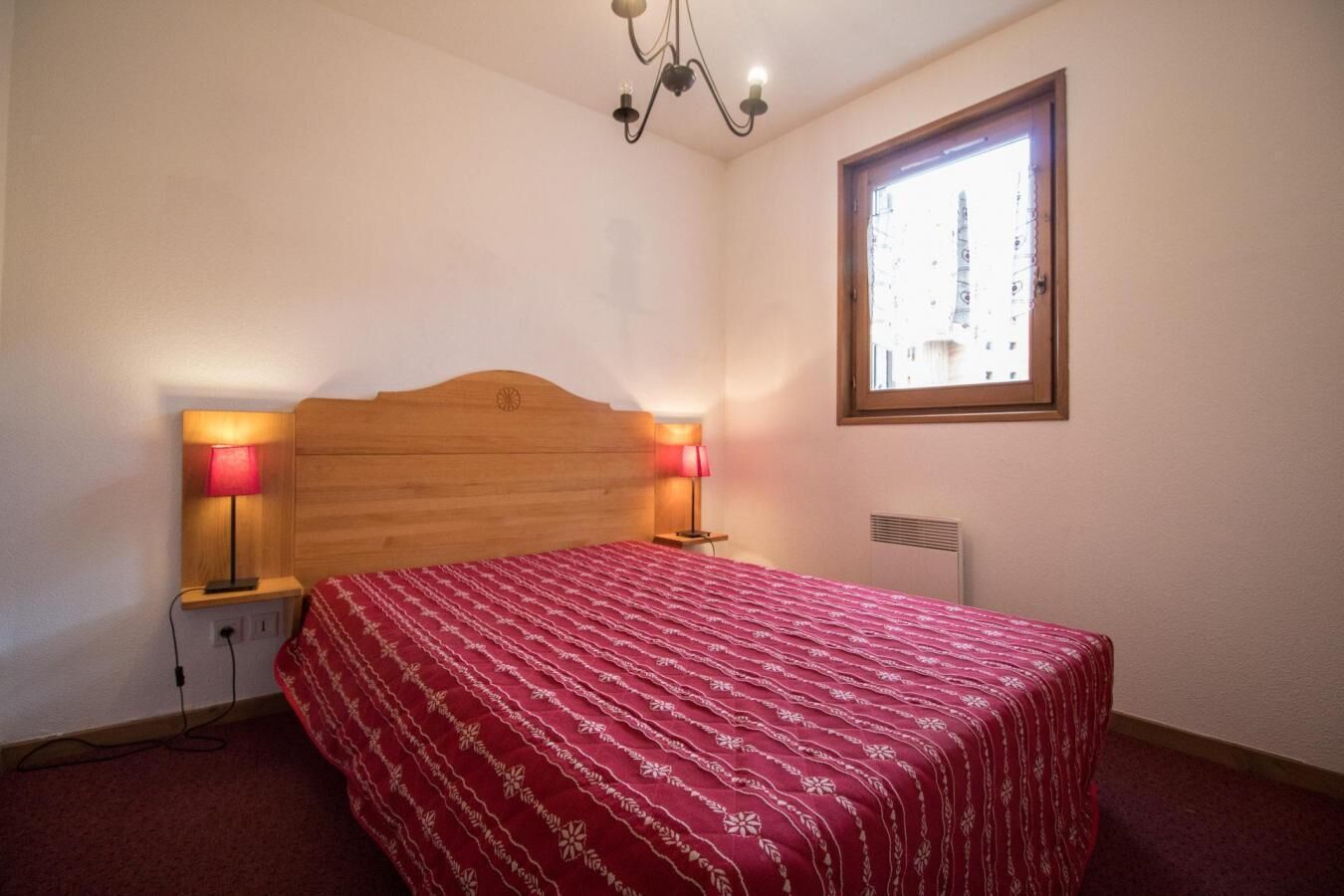 A bedroom with a wooden headboard, two bedside lamps, a window with a view, and a red patterned bedspread.