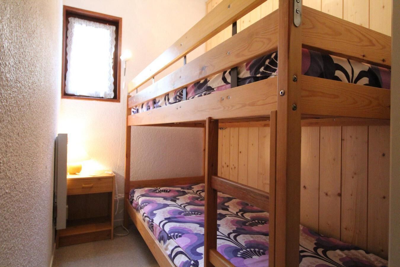 A bunk bed with a patterned bedspread, a bedside table with a lamp, and a window with a white curtain.