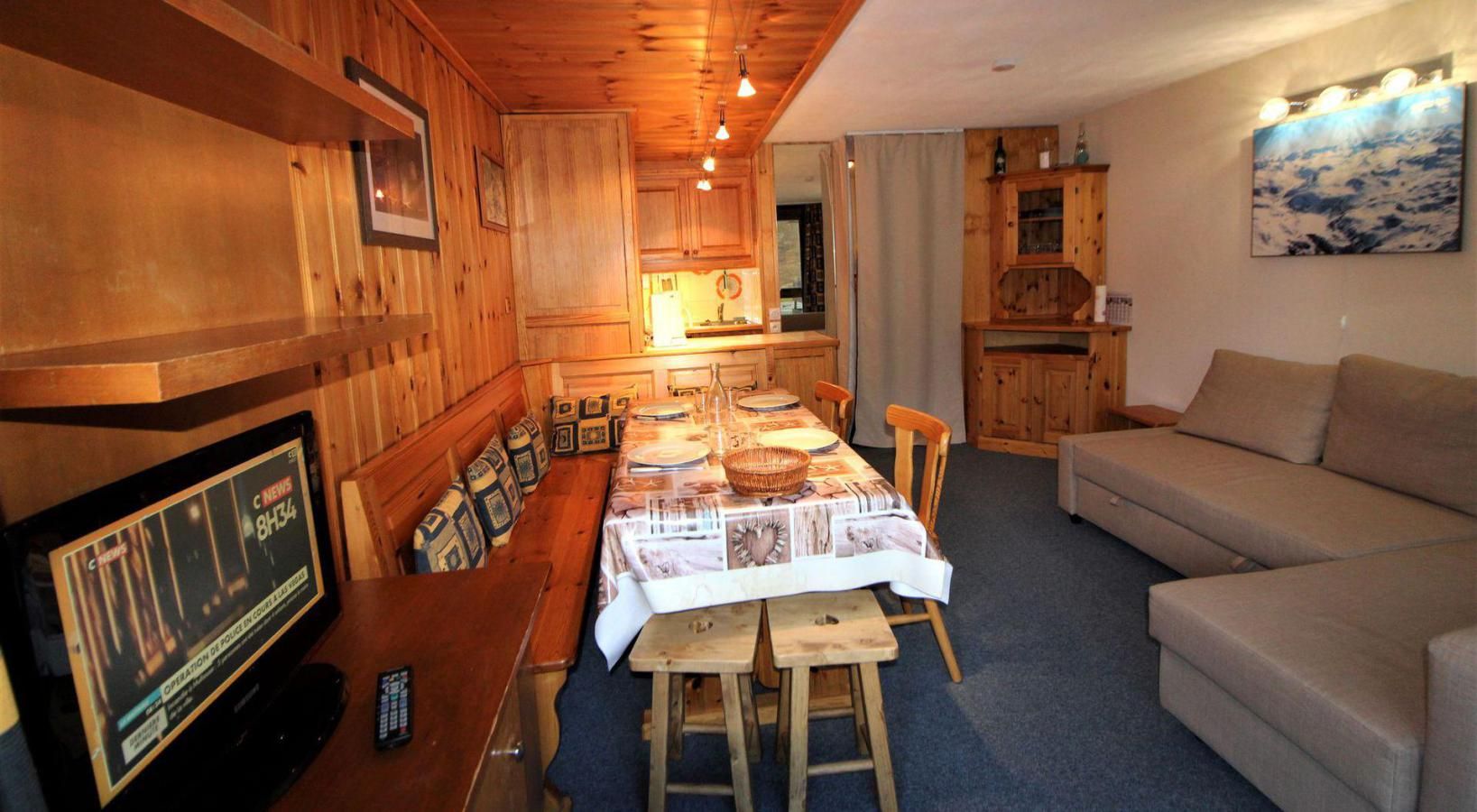 A cozy room with a wooden dining table, benches, a sofa, and a television displaying a news channel.