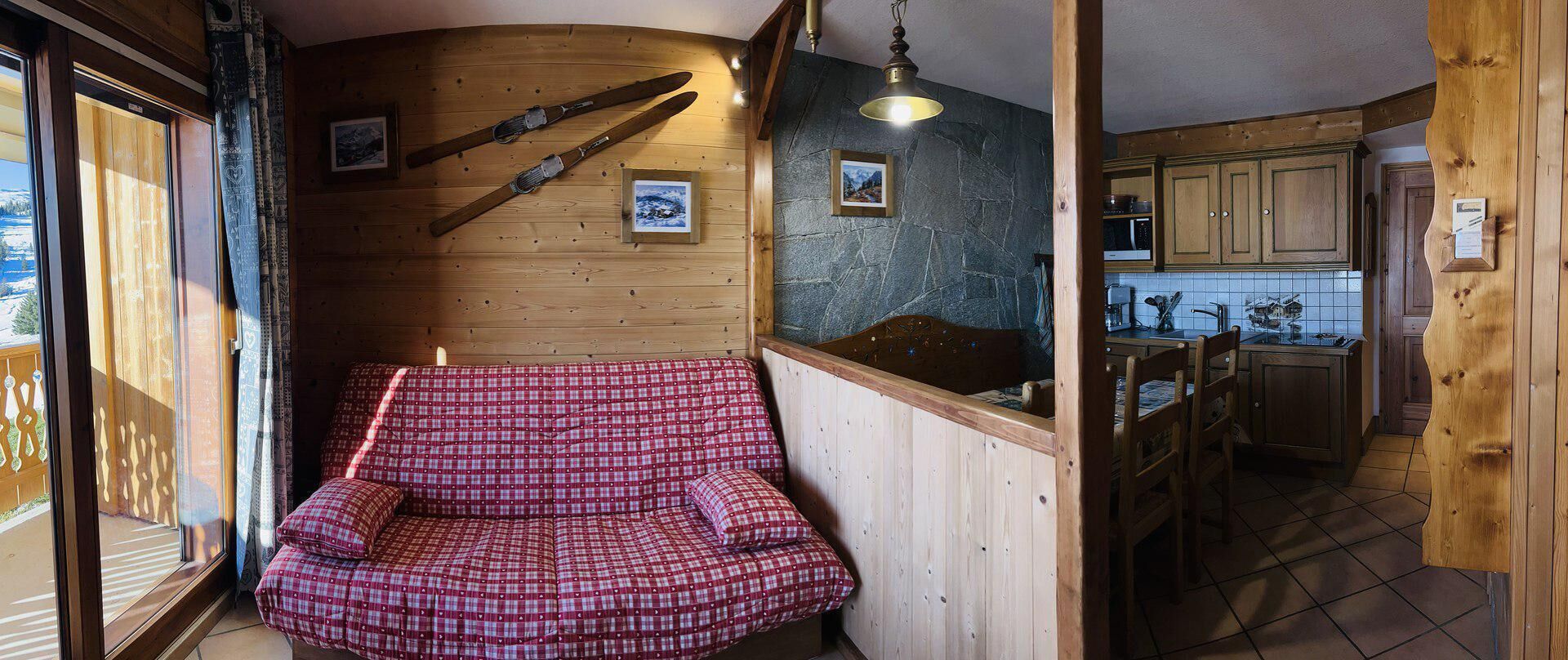 Une pièce chaleureuse avec un canapé en bois, un mur en pierre et une cuisine visible à travers une porte ouverte.