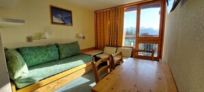 A room with a wooden sofa, a small table, and chairs. There is a window with curtains and a view of mountains.