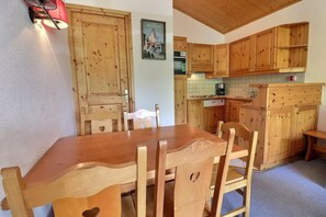 A wooden dining table with chairs, a kitchen with wooden cabinets, and a wall-mounted light fixture. - 10m from ski slopes, 4th floor, balcony, 40m², Méribel-Mottaret (Méribel - Les Allues)